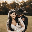 Bride Wearing Lacey Sheath Long Sleeve Wedding Dress Called Francesca By Maggie Sottero Held From Behind By Cowboy Hat Groom