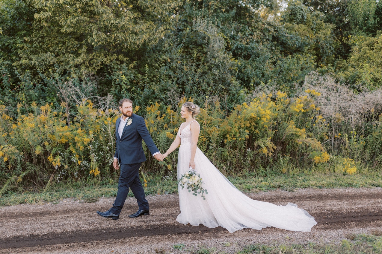 Bride wearing wedding dress called Pierce holding hands with groom walking along dirt path.