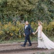 Bride wearing wedding dress called Pierce holding hands with groom walking along dirt path.