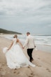 Bride wearing Indiana by Maggie Sottero walking on the beach with her husband