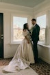 Bride Wearing Floral Ballgown Wedding Dress Called Derrick Marie Standing Next To Groom Next To Windows