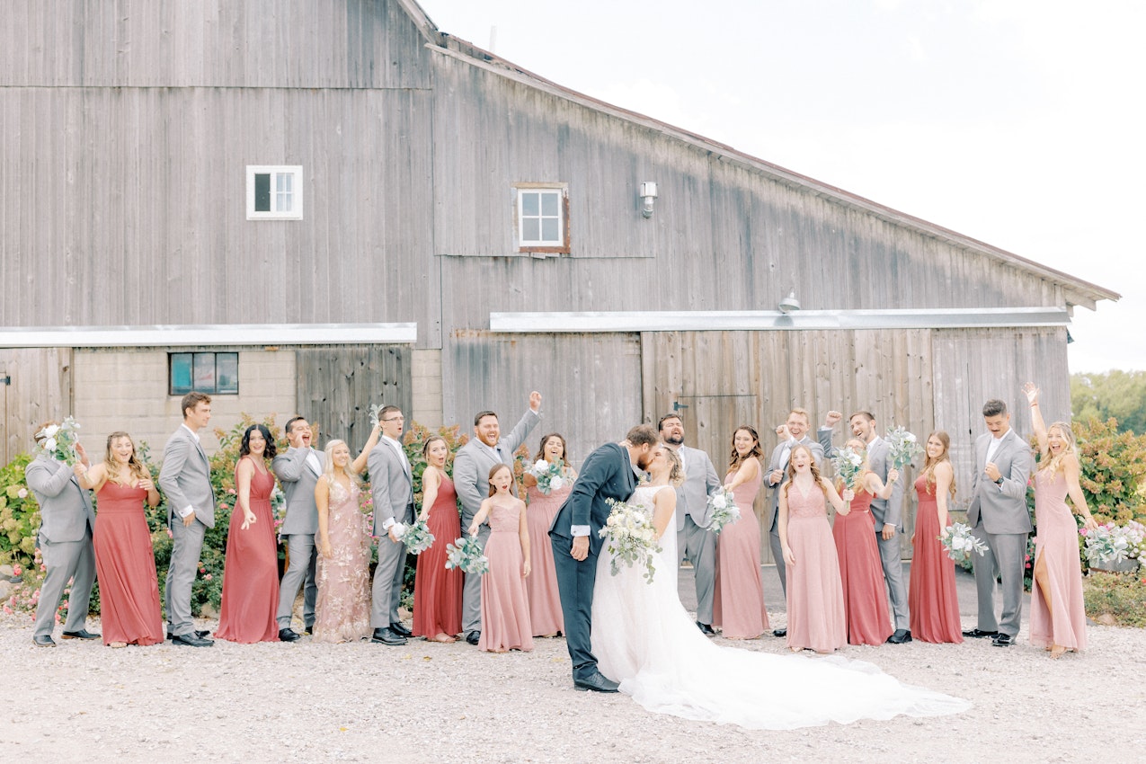 Wedding party in salmon and grey colors cheer as groom kisses his bride.