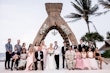 Wedding party with bride and groom stood under straw archway. 