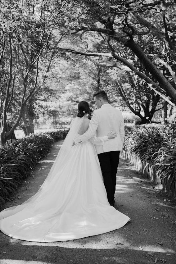 Newlyweds with arms around each other walking on a path surrounded by trees and bushes.