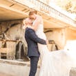 Groom Picks Up Bride Wearing Glamorous Lace And Sequin Wedding Dress Called Tuscany By Maggie Sottero Under Bridge