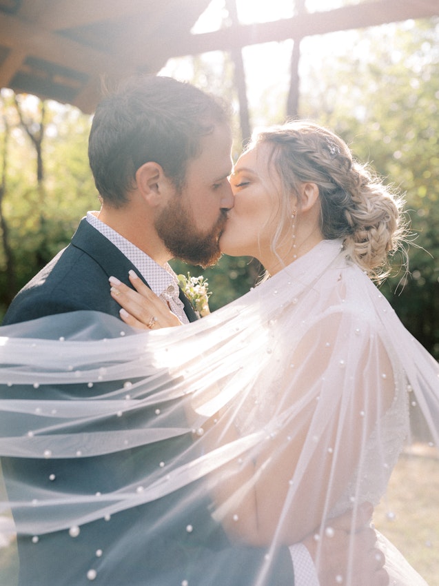 Bride and groom kissing while veil adorned with pearls cascades their shoulders.