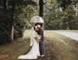 Bride Wearing Lacey Sheath Long Sleeve Wedding Dress Called Francesca By Maggie Sottero Kissing Groom Under Umbrella