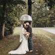 Bride Wearing Lacey Sheath Long Sleeve Wedding Dress Called Francesca By Maggie Sottero Kissing Groom Under Umbrella