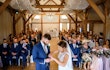Bride placing ring on her husband to be with wedding guests in the background.