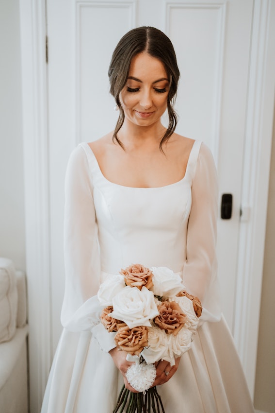 Bride holding champagne and ivory colored bouquet looking down at the flowers.