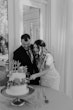 Groom Cutting Cake With Bride In Illusion V-back Sheath Wedding Dress Named Angie By Rebecca Ingram