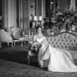 Bride Wearing Simple Off-the-shoulder Wedding Dress Called Josie By Rebecca Ingram Sitting On A Couch