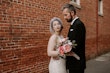 Bride Wearing Rustic Lace Wedding Dress Called Larkin Lynette By Rebecca Ingram With Groom Next To A Red Brick Wall
