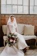 Bride Wearing Simple Fit And Flare Wedding Dress Called Josie By Rebecca Ingram Sitting On A Bench