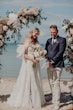 Bride Wearing Off-The-Shoulder Lace Wedding Dress Called Carson By Sottero And Midgley By A Flower Alter With Groom