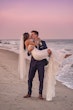 Groom Holding Bride on Sand Beach with Sunset Background Wearing Relaxed Boho Mermaid Wedding Dress called Tuscany by Maggie Sottero.