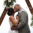 Bride Wearing Simple Chiffon Wedding Dress Called Marta Lynette By Rebecca Ingram Kissing Groom Under Triangle Arch With Flowers