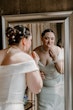 Bride Wearing Simple Fit And Flare Wedding Dress Called Josie By Rebecca Ingram Looking In A Mirror Putting On Makeup