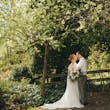 Bride Wearing Chiffon A-Line Wedding Dress Called Heather By Rebecca Ingram Kissing Groom In A Garden